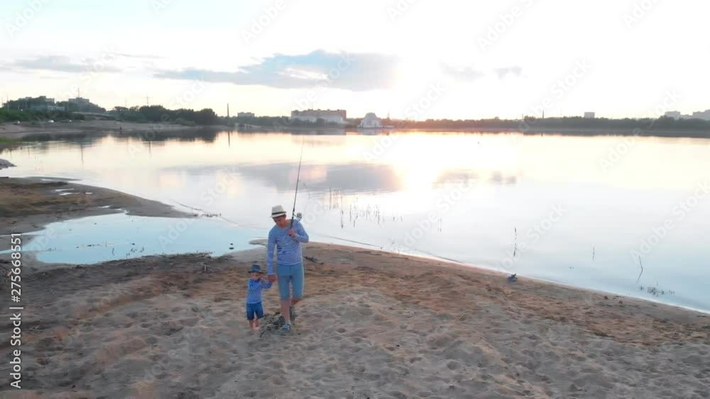 A father and his son in pair clothes walking on fishing on the river beach holding hands A father and his son in pair clothes walking on fishing on the river beach holding hands