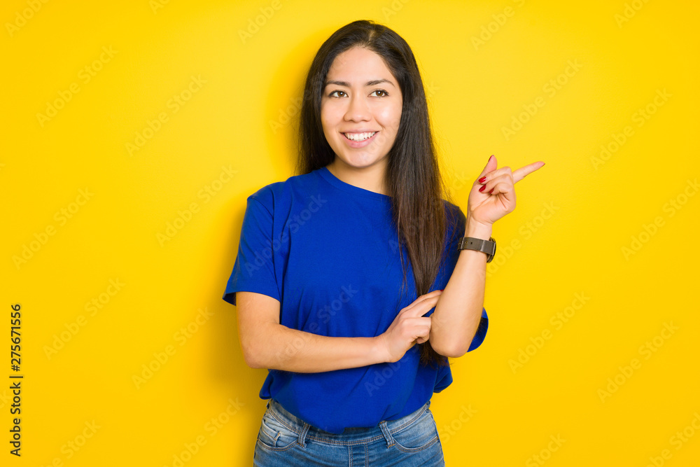 Beautiful brunette woman wearing blue t-shirt over yellow isolated background with a big smile on face, pointing with hand and finger to the side looking at the camera.