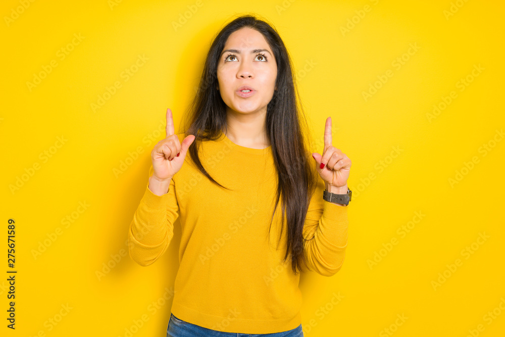 Beautiful brunette woman over yellow isolated background amazed and surprised looking up and pointing with fingers and raised arms. Beautiful brunette woman over yellow isolated background amazed and surprised looking up and pointing with fingers and raised arms.