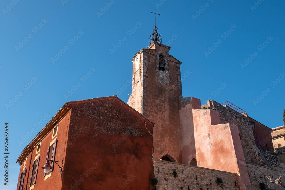 Roussillon, France: church tower and buildings facade with the ochre color with clean blue sky as background