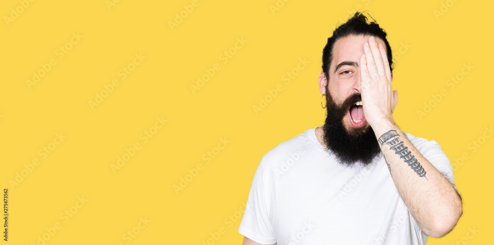 Young hipster man with long hair and beard wearing casual white t-shirt covering one eye with hand with confident smile on face and surprise emotion. Young hipster man with long hair and beard wearing casual white t-shirt covering one eye with hand with confident smile on face and surprise emotion.