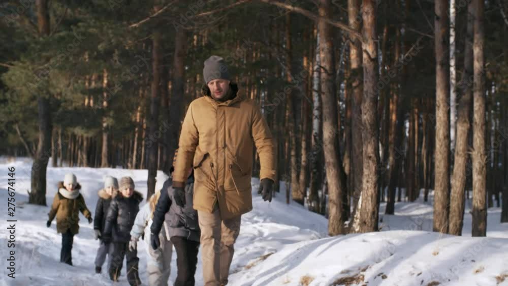 Full shot of middle-aged father with his five bubbly young kids, dressed in warm winter clothes, walking in single file along footpath in snow-covered pine forest, talking and smiling Full shot of middle-aged father with his five bubbly young kids, dressed in warm winter clothes, walking in single file along footpath in snow-covered pine forest, talking and smiling