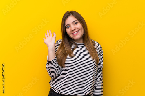 Young girl over yellow wall saluting with hand with happy expression