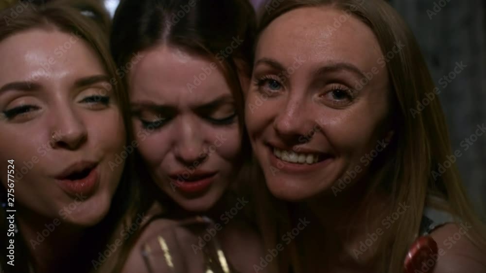 Close-up racking focus three shot of young beautiful 20 something partying girls posing for the camera in dark room, pulling funny faces, blowing kisses and toasting with glass of champagne