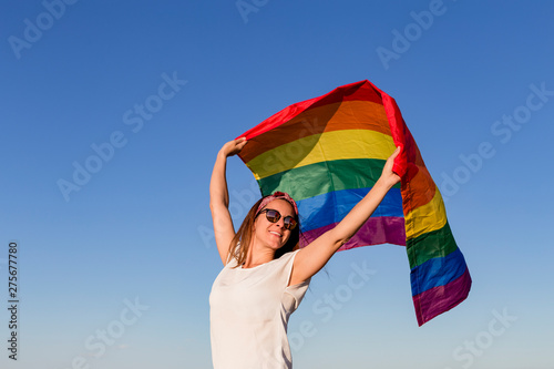 Woman holding the Gay Rainbow Flag over blue sky outdoors. Happiness, freedom and love concept for same sex couples. LIfestyle outdoors
