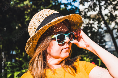 Young woman wearing summer straw hat and sunglasses sitting in the sun while looking in the distance – Casually dressed girl arranging her eyeglasses outdoor