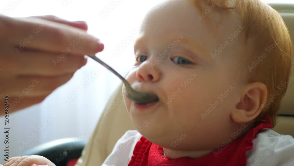 Baby eating puree with spoon. Close up of infant baby face eat baby food. Mother feeding child with porridge. Little girl eating porridge Baby eating puree with spoon. Close up of infant baby face eat baby food. Mother feeding child with porridge. Little girl eating porridge
