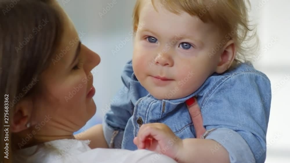 Tilting close-up shot of young Caucasian mother holding up her blond blue-eyed baby boy, gently speaking to him and blowing softly at his hair, while boy is smiling and staring away
