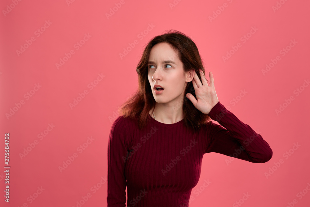 Surprised young lady standing isolated over dark pink background with hands raised close to ear as if is listening to some rumors. Surprised young lady standing isolated over dark pink background with hands raised close to ear as if is listening to some rumors.