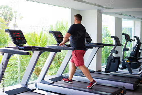 chubby man walking on running track, warming up on gym treadmill. back view
