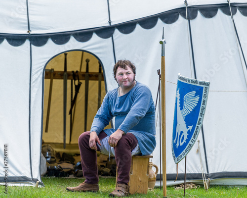 A medieval armorer sits outside his tent.
