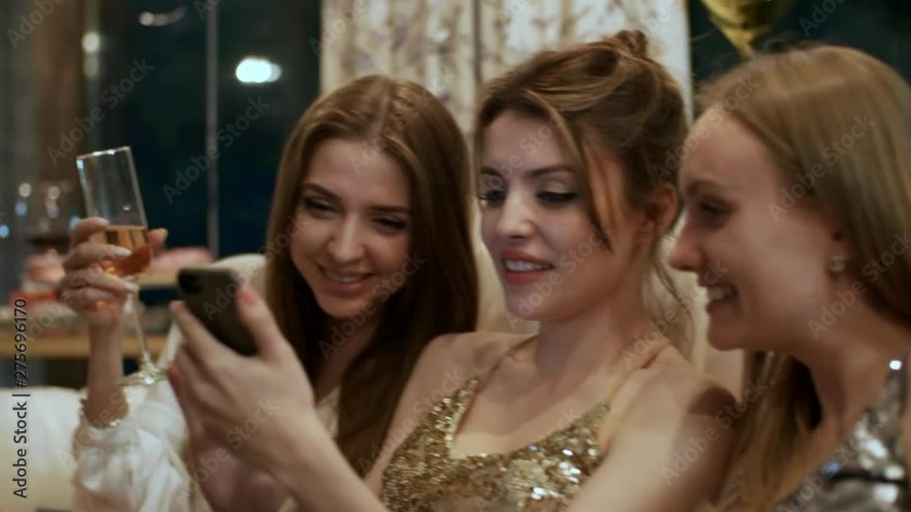 Medium shot of three sexy relaxed Caucasian girlfriends sitting together at party, checking updates on smartphone and laughing animatedly Medium shot of three sexy relaxed Caucasian girlfriends sitting together at party, checking updates on smartphone and laughing animatedly