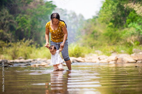 Fototapeta Naklejka Na Ścianę i Meble -  Asian woman holding her baby girl's hands to practice walking in water with beautiful forest view in background