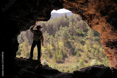 MOUNTAIN MEN ADVENTURER WITH AUSTRALIAN HAT AND BACKPACK IN A CAVE WITH A PINE FOREST IN THE BACKGROUND