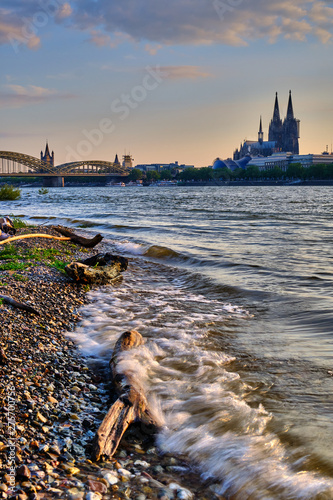 Köln Stadtportrait bei Sonnenuntergang, starken Wellen im Vordergrund, Kölner Dom und Hohenzollernbrücke im Hintergrund. Kölner Skyline Poster und Postkartenmotiv im Hochformat.