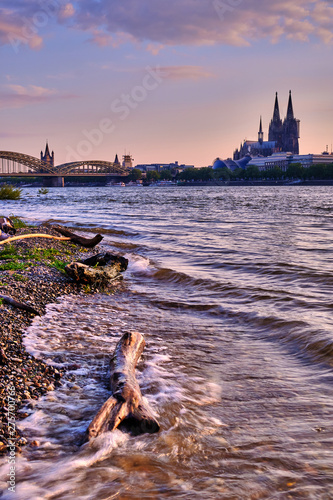 Köln Stadtportrait bei Sonnenuntergang, starken Wellen im Vordergrund, Kölner Dom und Hohenzollernbrücke im Hintergrund. Kölner Skyline Poster und Postkartenmotiv im Hochformat.