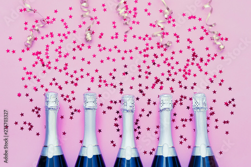 Five Champagne bottles with confetti stars and party streamers on pink background. Copy space,top view. Party background photo