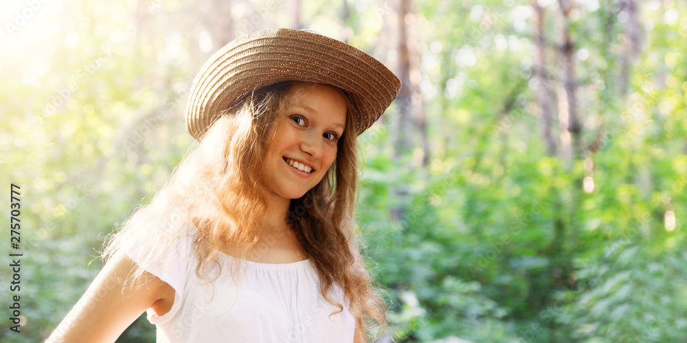 Closeup portrait of a young longhair girl in a straw hat in bright sunlight against blurred nature background. Copyspace.