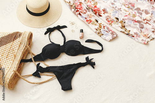 Black bikini swimsuit, floral shirt dress, straw boater hat, wicker beach bag, gold rings on beige background. Woman's swimwear and beach accessories. Flat lay, top view. Beach outfit.