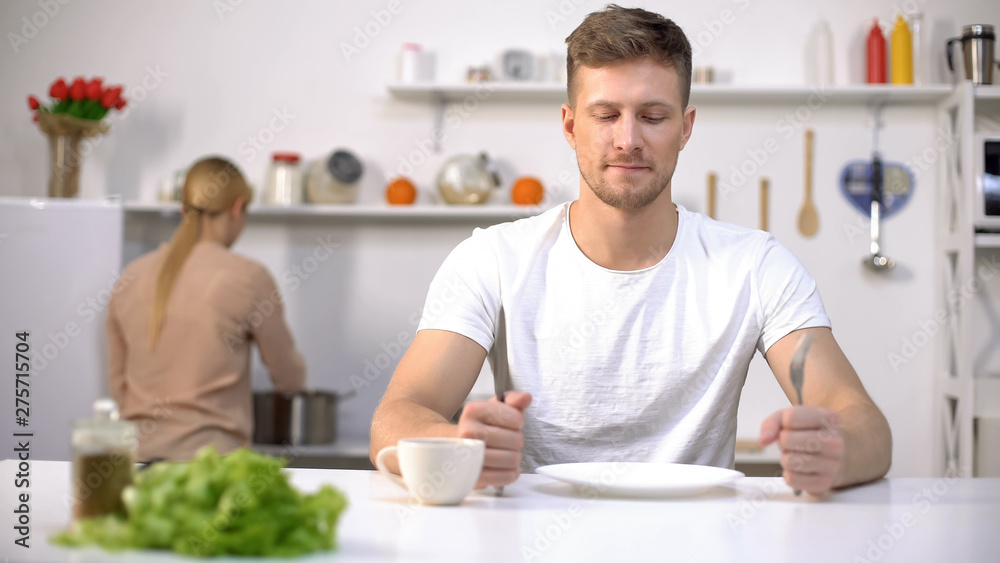 Positive man waiting for dinner, young wife cooking soup on background, hunger Positive man waiting for dinner, young wife cooking soup on background, hunger