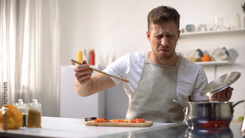 Fototapeta premium Young man tasting cooked food with disgusted face expression, funny grimacing