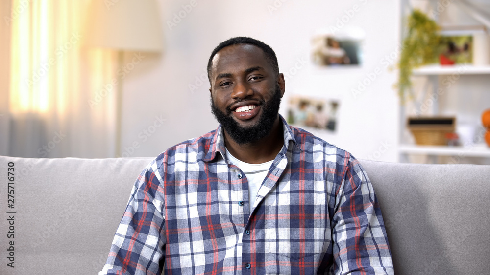 Obraz premium Cheerful African-American man sitting on sofa and looking at camera, relaxing