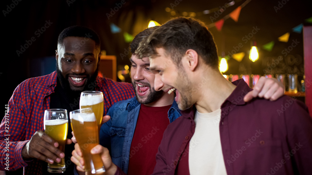 Joyful multiethnic bachelors clinking beer glasses, watching football in pub