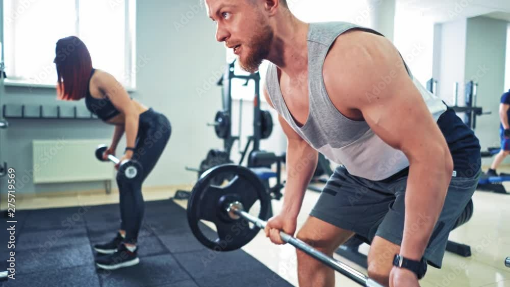 Athletic couple in sportswear lifting dumbbells in the gym. Persistent man is doing hard workout with dumbbells standing in special pose in the sports club.