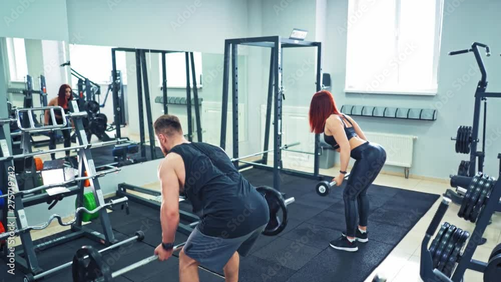 Back view of athletic man and woman lifting barbells in the modern gym. Strong couple is doing physical exercises with heavy equipment standing in front of a big mirror in fitness club. Back view of athletic man and woman lifting barbells in the modern gym. Strong couple is doing physical exercises with heavy equipment standing in front of a big mirror in fitness club.