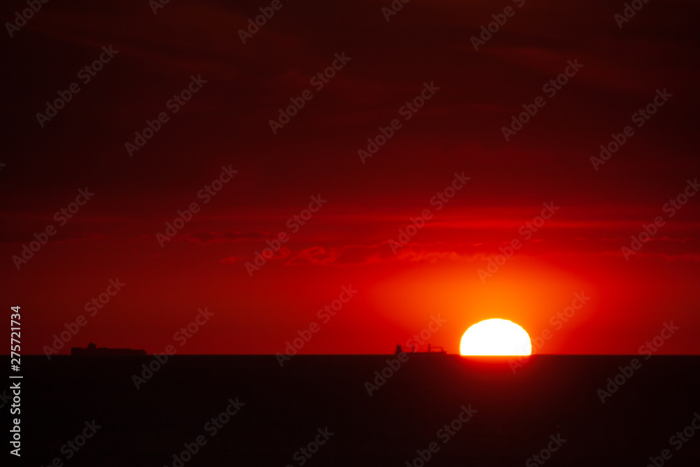  Sunset over the sea at the harbor entrance to Rotterdam. The sun touches the sea. Silhouette of ships in front of glowing red background.