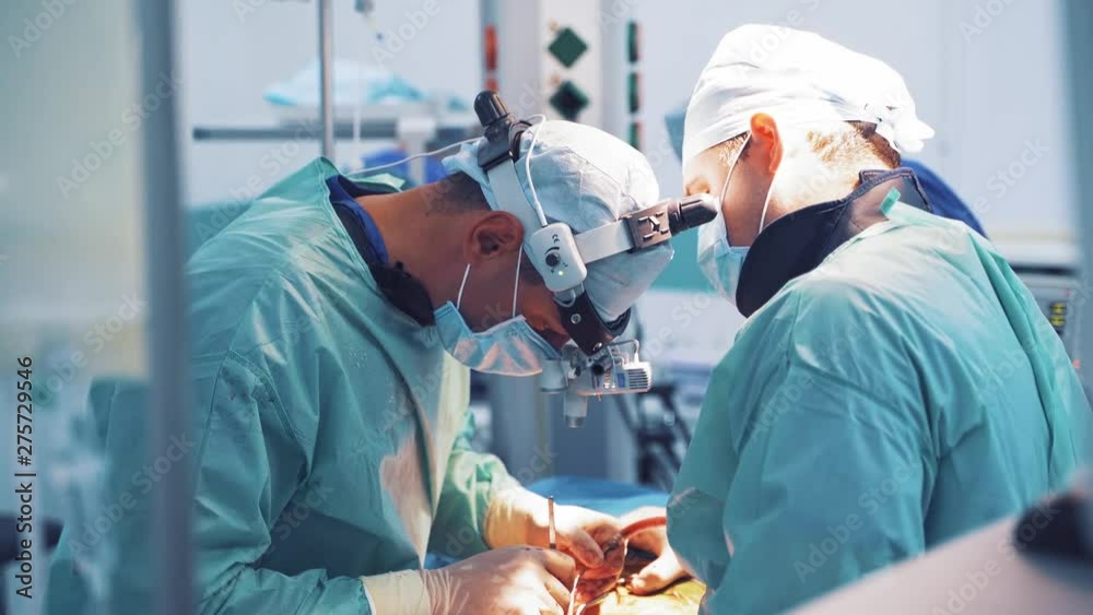 Surgeon with magnifying glass on his head is working with assistants in operation room. Team of specialists in medical uniform performing an operation to a patient with special instruments in clinic.
