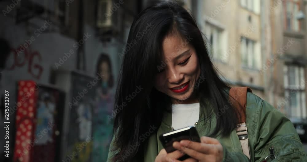 Attractive young Asian woman in stylish outfit chatting and typing on the smartphone with interest and smile at the street with graffity. Close up.