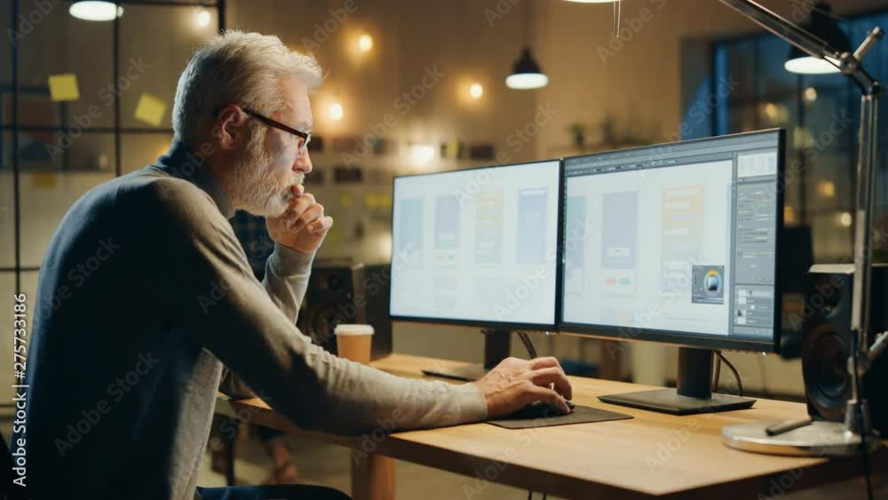 Creative Middle Aged Mobile Software Designer Sitting at His Desk Uses Desktop Computer with Two Screens Showing Smartphone Application Design Process. Stylish Modern Studio Office Creative Middle Aged Mobile Software Designer Sitting at His Desk Uses Desktop Computer with Two Screens Showing Smartphone Application Design Process. Stylish Modern Studio Office