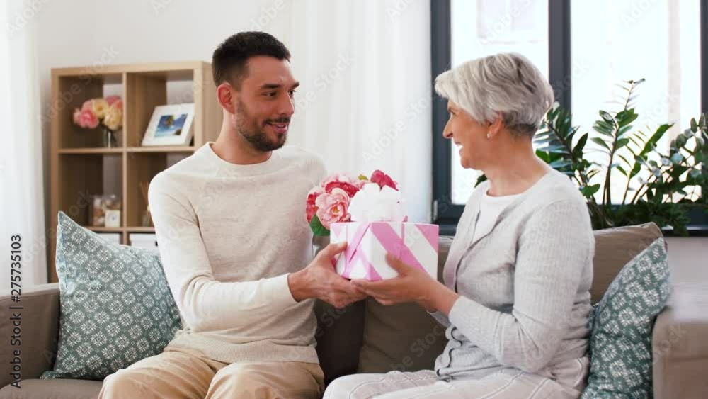 family, mother's day and birthday concept - smiling adult son giving present and flowers to his senior mother at home