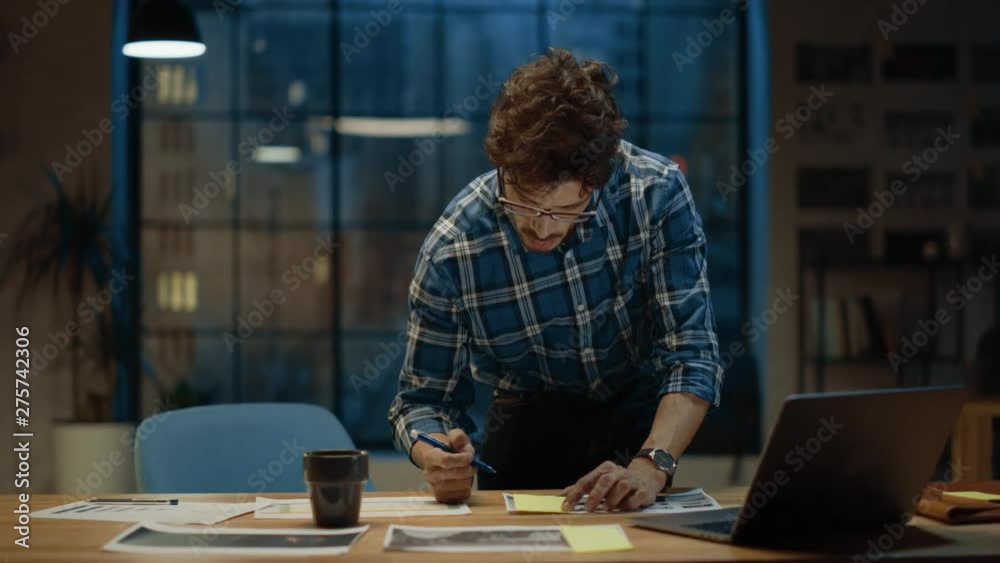 Young Creative Designer Working at His Desk, Concentrated on the Paperwork, Choosing Right Design, Concept and Sketches. He Drinks from a Cup. In the Background Evening Office Studio with Window Young Creative Designer Working at His Desk, Concentrated on the Paperwork, Choosing Right Design, Concept and Sketches. He Drinks from a Cup. In the Background Evening Office Studio with Window