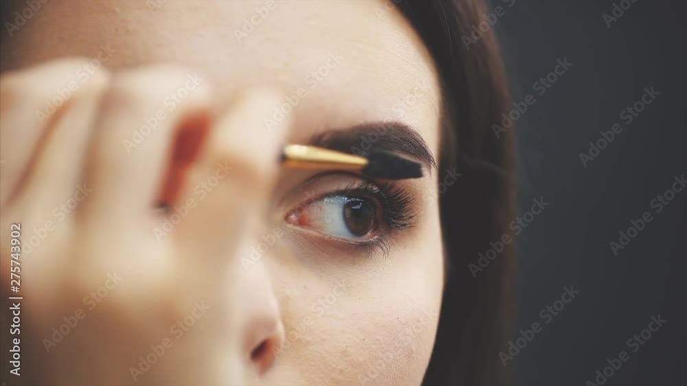 Beautiful young girl stands on a black background. During this, looking a mirror and using a brush makes an eyebrow makeup. Beautiful young girl stands on a black background. During this, looking a mirror and using a brush makes an eyebrow makeup.