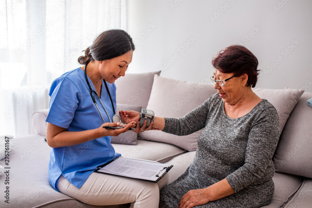 Happy senior woman having her blood pressure measured in a nursing home by her caregiver. Happy nurse measuring blood pressure of a senior woman in living room Happy senior woman having her blood pressure measured in a nursing home by her caregiver. Happy nurse measuring blood pressure of a senior woman in living room