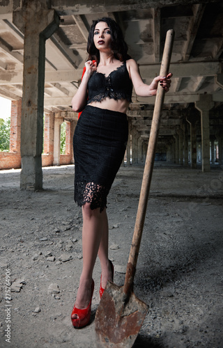 Portrait of beautiful girl with shovel in hand. Elegant young woman dressed in black skirt and brassiere among abandoned industrial place © jetrel2