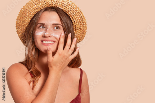 Photo of attractive smiling woman with long hair, has happy facial expression, applaying sunscreen, wearing straw hat, wanting to tan, isolated on beige wall. Summertime, vacation, sunscreen concept. photo