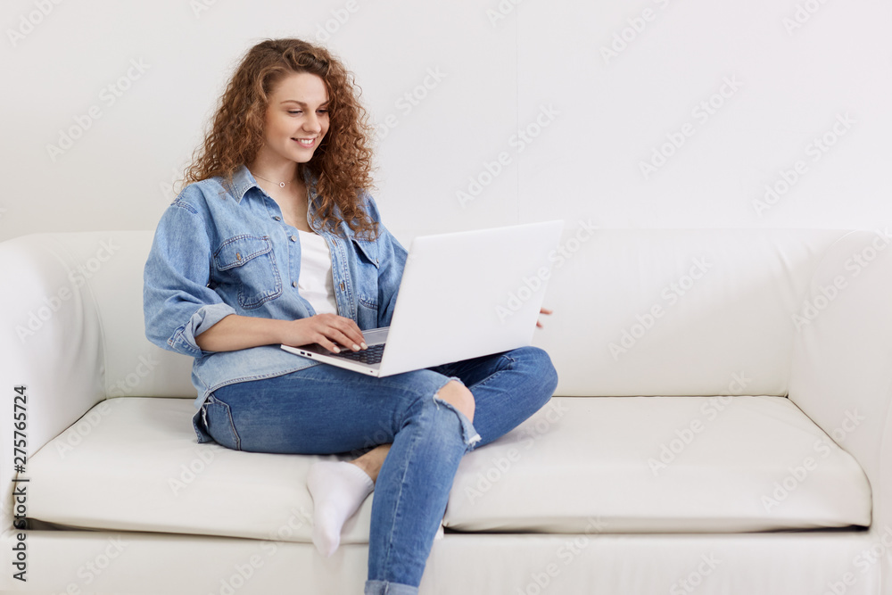 Horizontal shot of nice looking lovely attractive woman, focused curly haired girl sitting on white sofa while working remotely part time, using her modern portable computer and wireless Internet. Horizontal shot of nice looking lovely attractive woman, focused curly haired girl sitting on white sofa while working remotely part time, using her modern portable computer and wireless Internet.