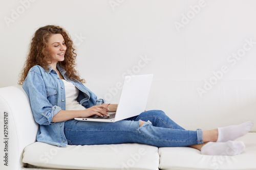 Happy cheerful young female lying on her white sofa, having rest, holding laptop on her legs, looking at screen attentively, typing information, surfing internet, being in good mood. Free time.