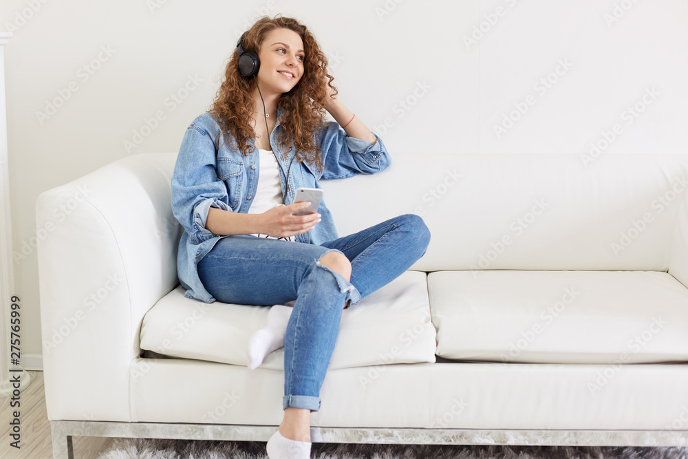 Cheerful pleasant female sitting on sofa, looking aside, holding her smartphone, having earphones, listening to favourite music, spending free time alone, wearing jeans clothes. Chill out concept.