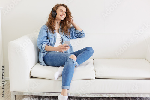 Free Download Cheerful pleasant female sitting on sofa, looking aside, holding her smartphone, having earphones, listening to favourite music, spending free time alone, wearing jeans clothes. Chill out concept. - GFXLoad