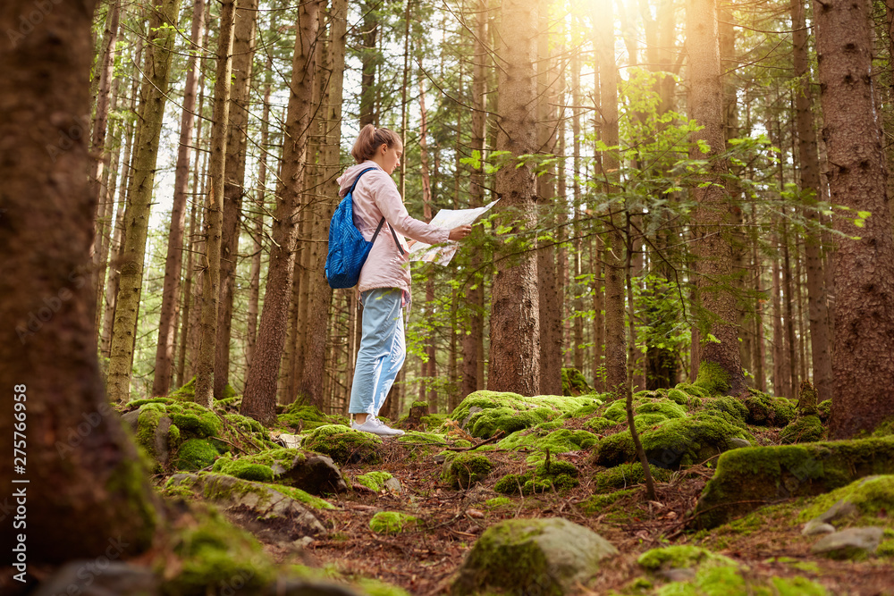 Profile of European slender active woman wearing rose jacket, jeans and blue backpack, holding map in both hands, searching for right way out of forest, go hiking, enjoying vacation. Orientating. Profile of European slender active woman wearing rose jacket, jeans and blue backpack, holding map in both hands, searching for right way out of forest, go hiking, enjoying vacation. Orientating.