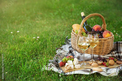 Fototapeta Naklejka Na Ścianę i Meble -  Picnic setting with  two glasses of white wine,  cheese, fruits and basket. Outdoor, green lawn