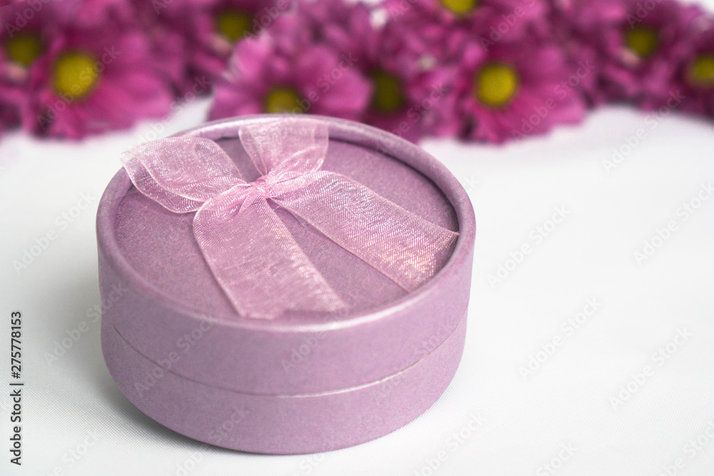 Purple round shape gift box with pink bow on white background and magenta flowers behind gift box. For design of cards for Valentine's day or Mother's day, invitations, wedding design. Copyspace Purple round shape gift box with pink bow on white background and magenta flowers behind gift box. For design of cards for Valentine's day or Mother's day, invitations, wedding design. Copyspace