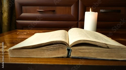 The old book lies on the antique table. The candle burns next to the book on the table.