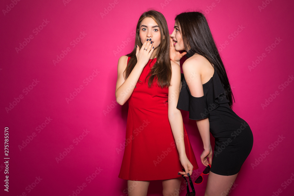 Two excited young girls dressed in summer clothes whispering secrets isolated over crimson background Two excited young girls dressed in summer clothes whispering secrets isolated over crimson background