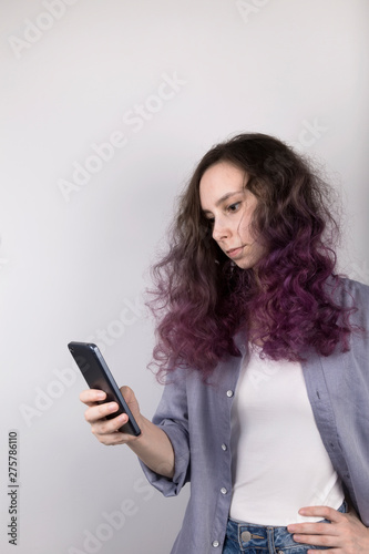 Young girl thoughtfully looks in phone. Curly color coloring purple hair. Gray background