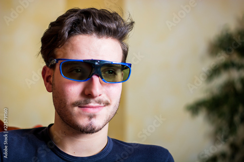 Young man in 3d glasses sitting on a sofa in the living room watching reality television leaning forwards with an absorbed expression